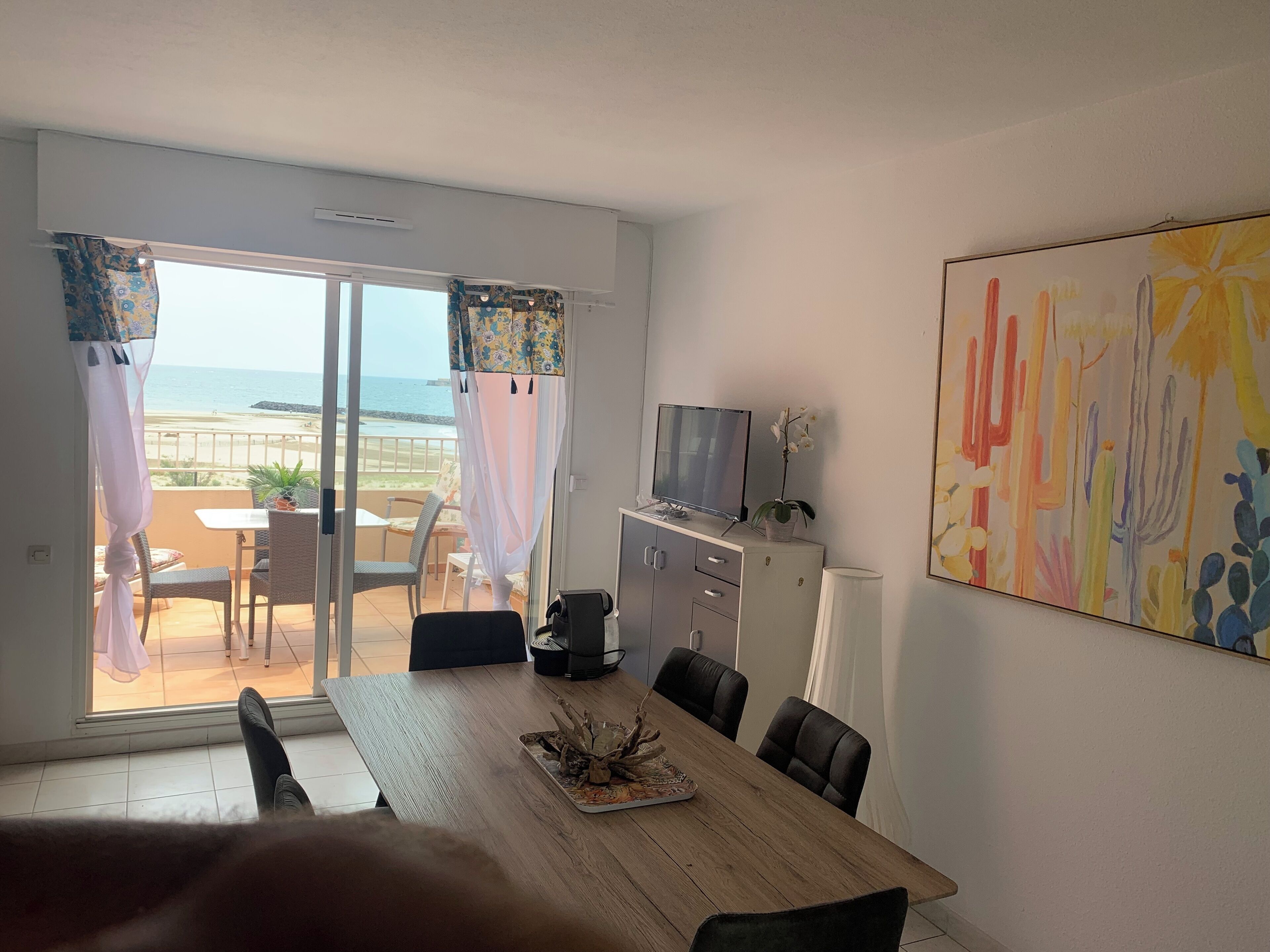 A room with a dining table, chairs, a TV on a cabinet, and a view of the beach through the window.