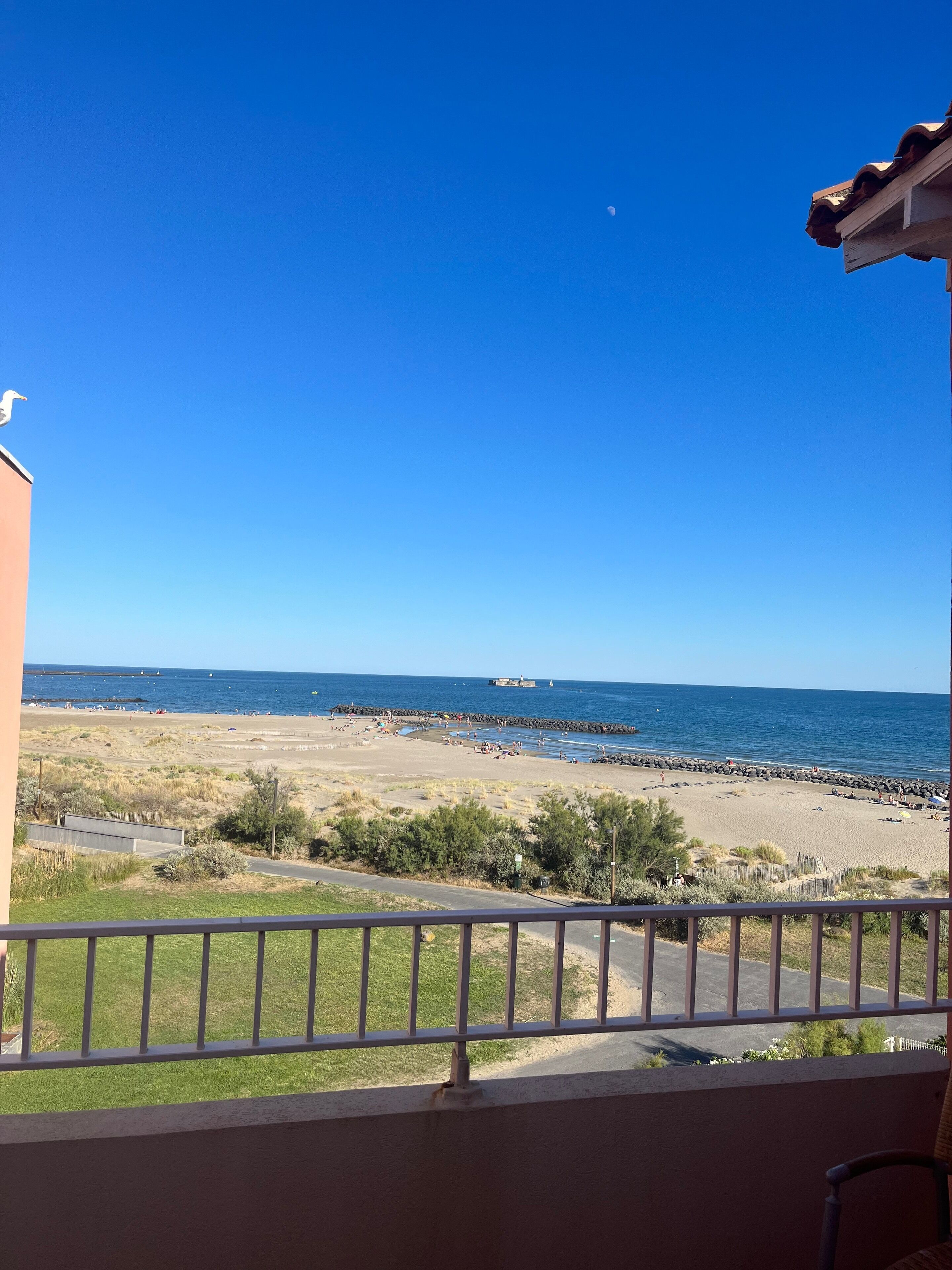 Un balcon donnant sur une plage, un ciel dégagé et un navire au loin.