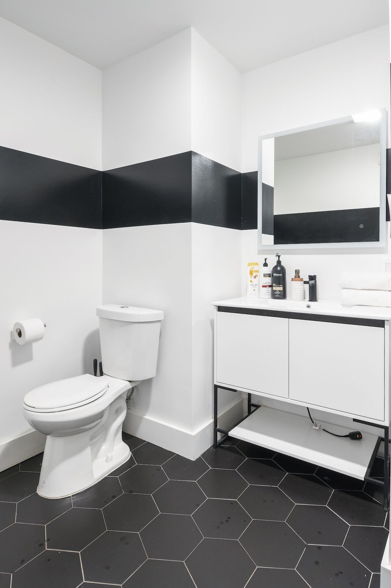 Une salle de bain moderne avec des toilettes blanches, des murs rayés de noir et de blanc, un lavabo blanc avec un miroir et des carreaux hexagonaux noirs sur le sol.