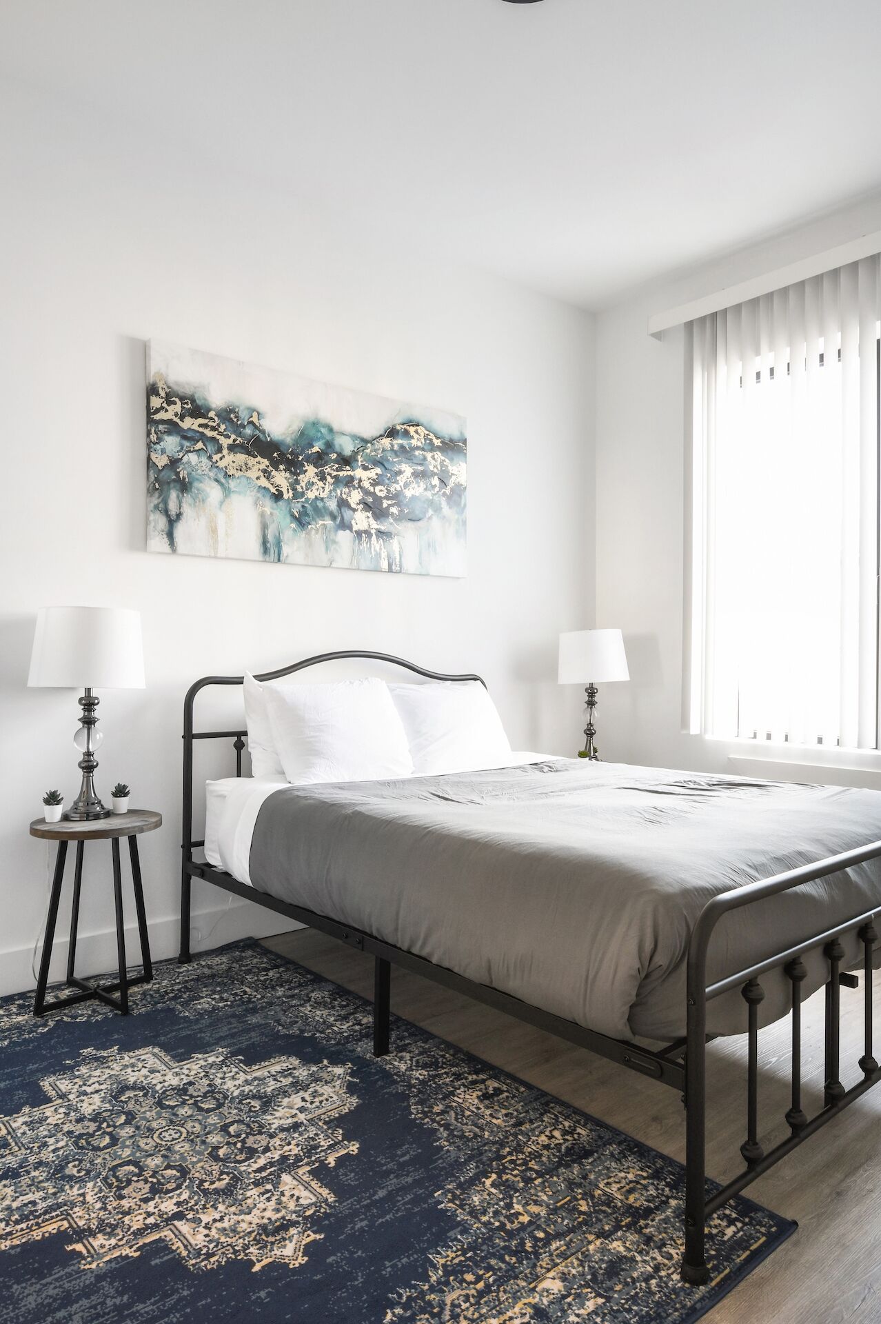 Een moderne slaapkamer met een metalen bed, een groot abstract schilderij, twee bedlampen en een vloerkleed met dessin.