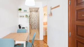 A dining area with a wooden table and blue chairs, a white pendant light, and a kitchen visible through an open door.
