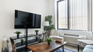 A modern living room with a flat-screen TV, a wooden coffee table, a sofa, and a window with blinds. - Cute 1BR Next to Airport cvts (Elizabeth)