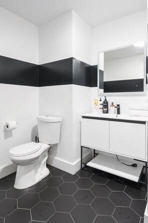 Une salle de bain moderne avec des toilettes blanches, des murs rayés de noir et de blanc, un lavabo blanc avec un miroir et des carreaux hexagonaux noirs sur le sol.