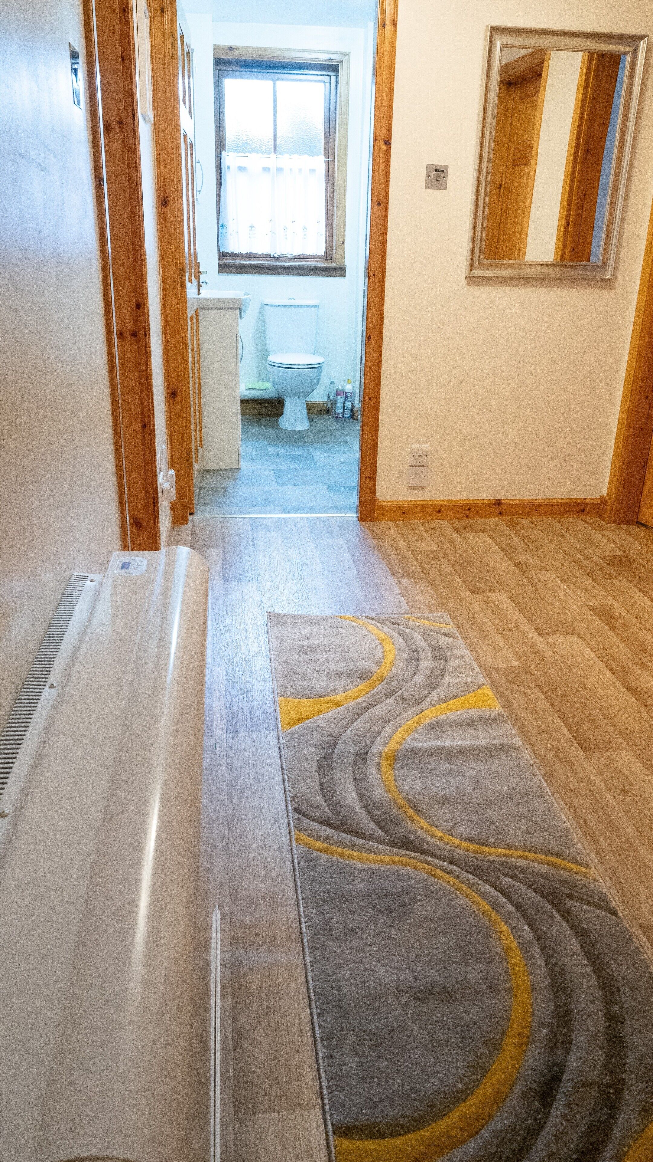 Un pasillo con piso de madera, alfombra gris con diseños amarillos y vistas a un baño con inodoro y ventana.