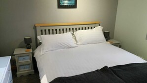 A neatly made bed with white linens, a wooden headboard, and two bedside lamps. There is a small bedside table with drawers and a lamp. A framed picture hangs on the wall above the bed. - Apartment 65B (Thurso)