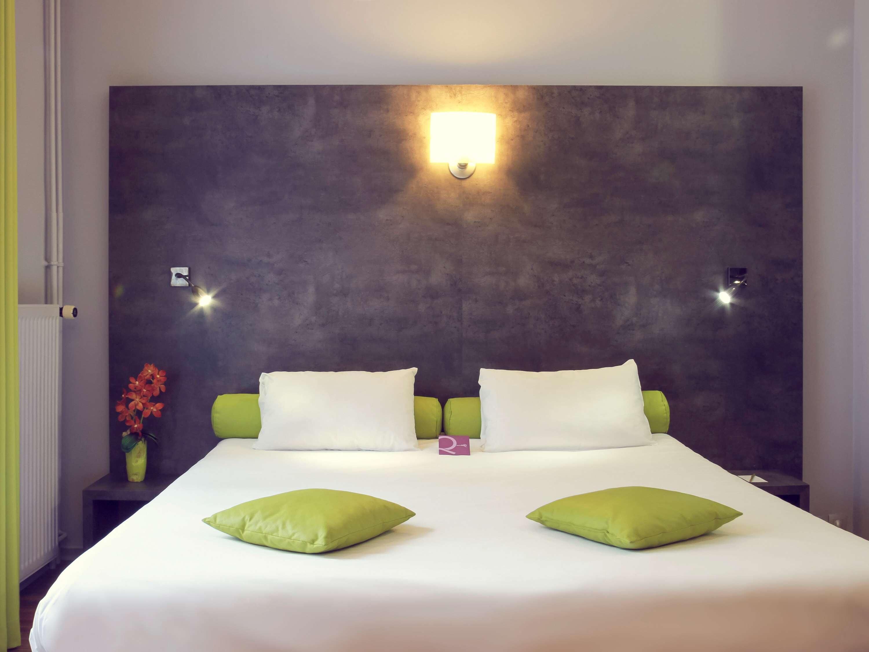 Habitació d'hotel amb un llit gran, dos coixins, una tauleta de nit, un vas amb flors i una làmpada penjant a la paret.