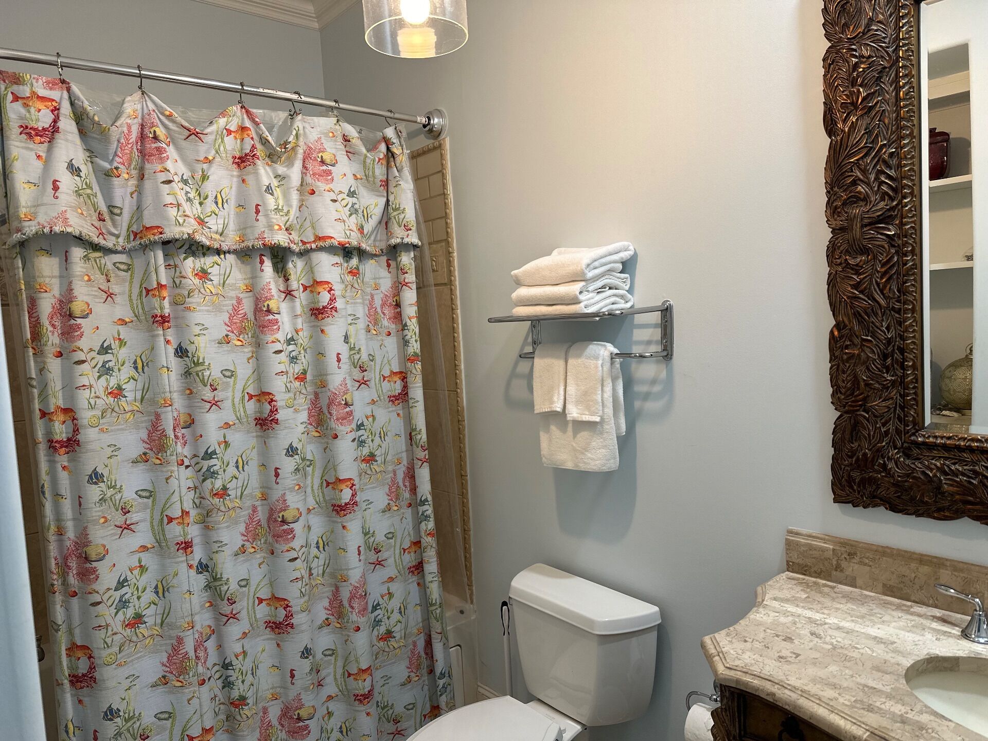 Une salle de bain avec un rideau de douche à motifs floraux, des toilettes, un porte-serviettes avec des serviettes pliées, un lavabo avec un plan-vasque en marbre et un miroir décoratif doté d’un cadre orné.