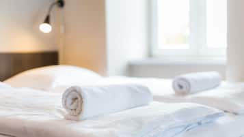 Cama bem arrumada com lençóis brancos e uma toalha enrolada, luminária de cabeceira e janela com luz do dia.