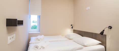 Una cama bien tendida con ropa de cama y toallas blancas, una mesita de noche con una lámpara y una ventana con vistas a zonas verdes.