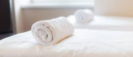 A neatly made bed with a rolled white towel on it, a white pillow, and a white blanket.