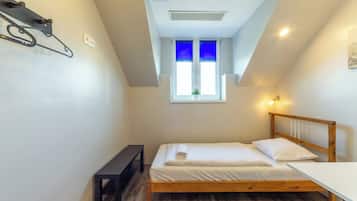 A small, neatly arranged bedroom with a single bed, a bedside table, a bench, and a window with blue blinds.