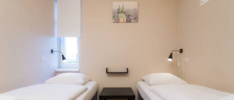 A small, clean room with two single beds, a small table, and a wall-mounted shelf.
