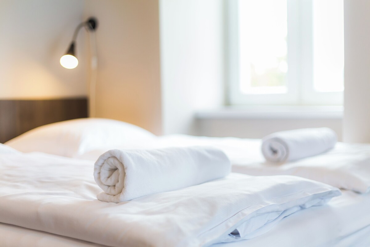 A neatly made bed with white linens and a rolled towel, a bedside lamp, and a window with daylight.