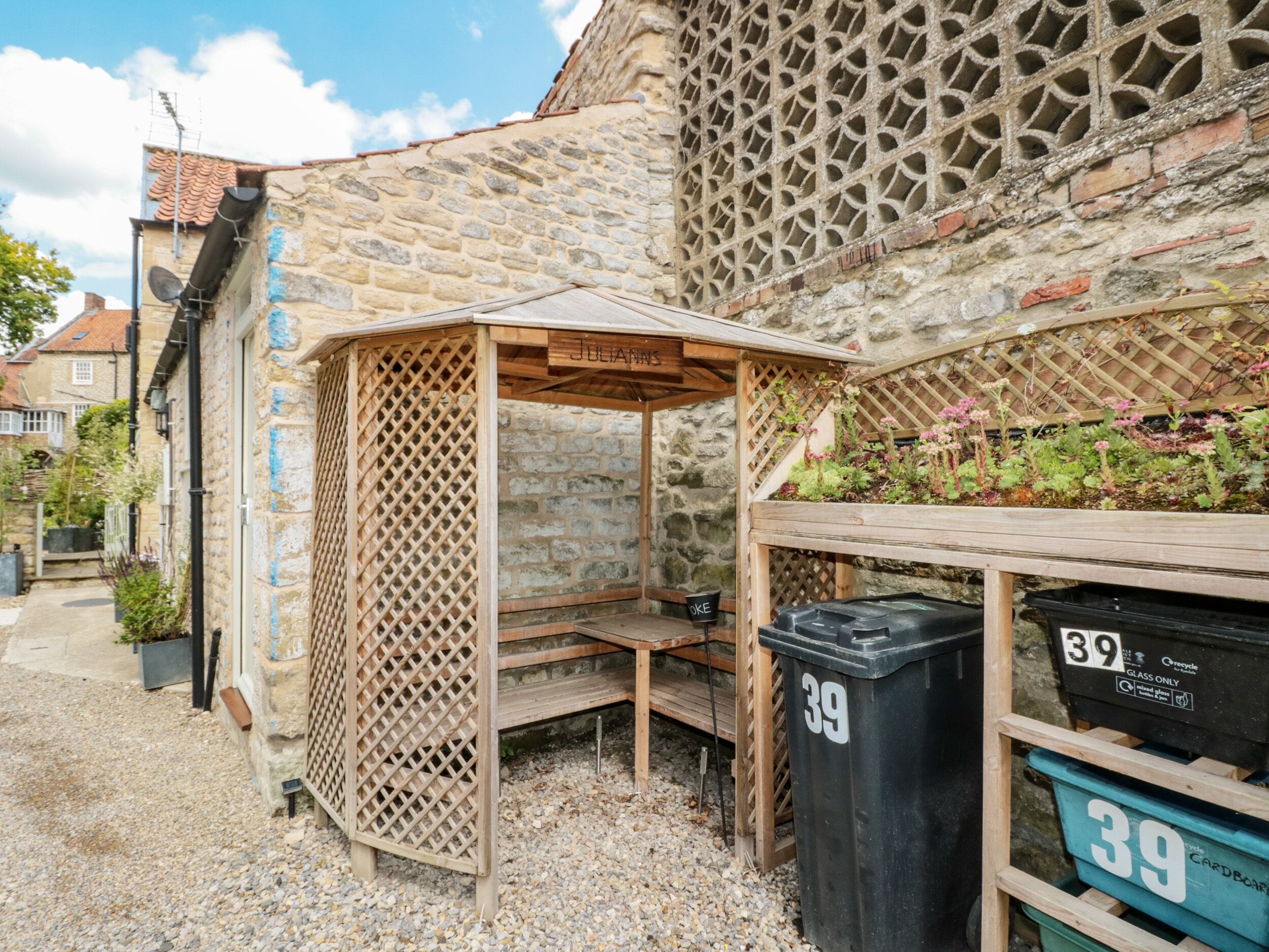 A stone building with a wooden bench and a small garden area. There are recycling bins labeled '39' and a wooden structure with a lattice design.
