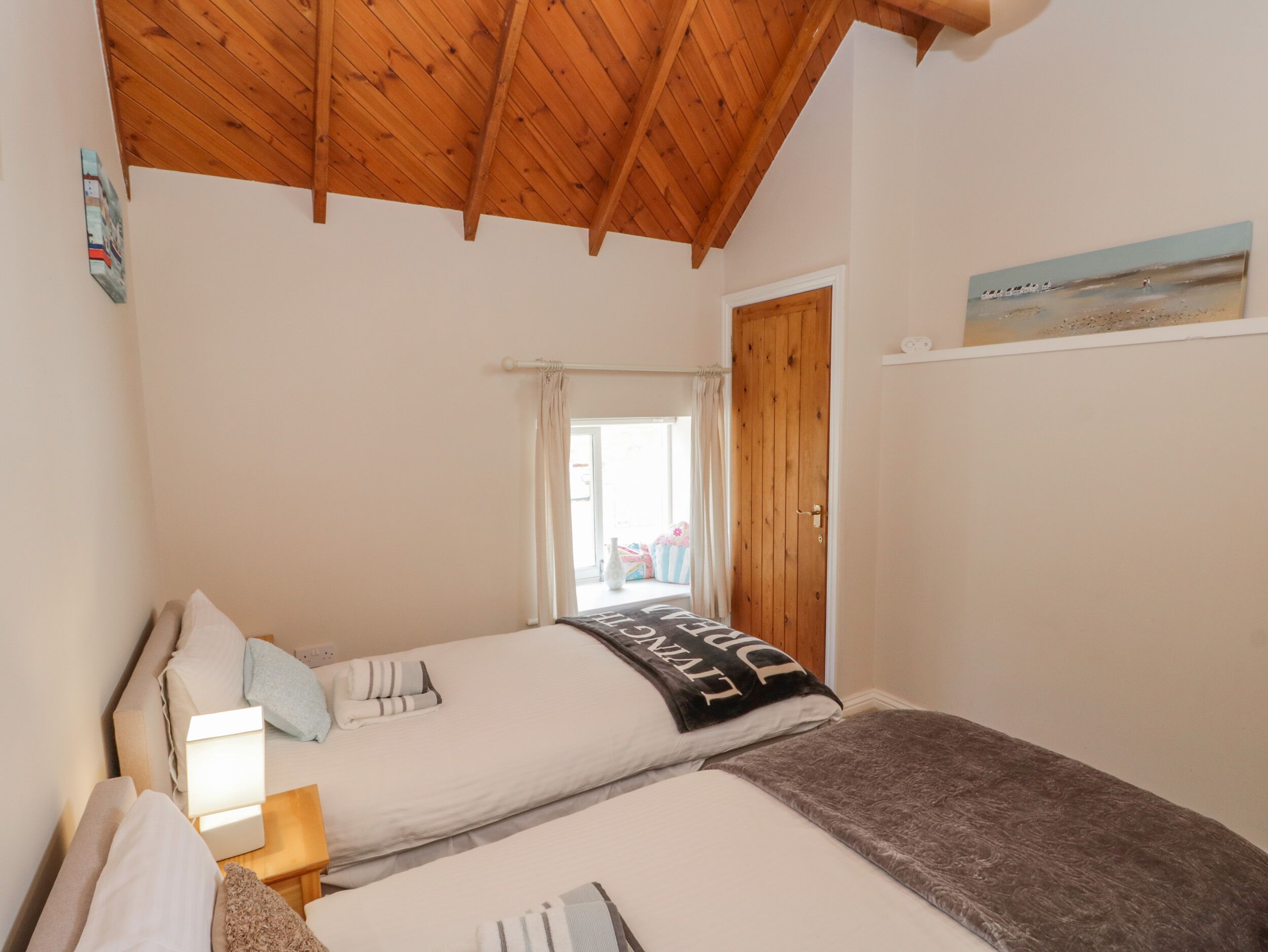 A bedroom with two beds, wooden ceiling beams, a window with curtains, and a door.