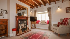A cozy living room with a fireplace, a television, a sofa, and a window with curtains. - Adelaide's Cottage (Pickering)