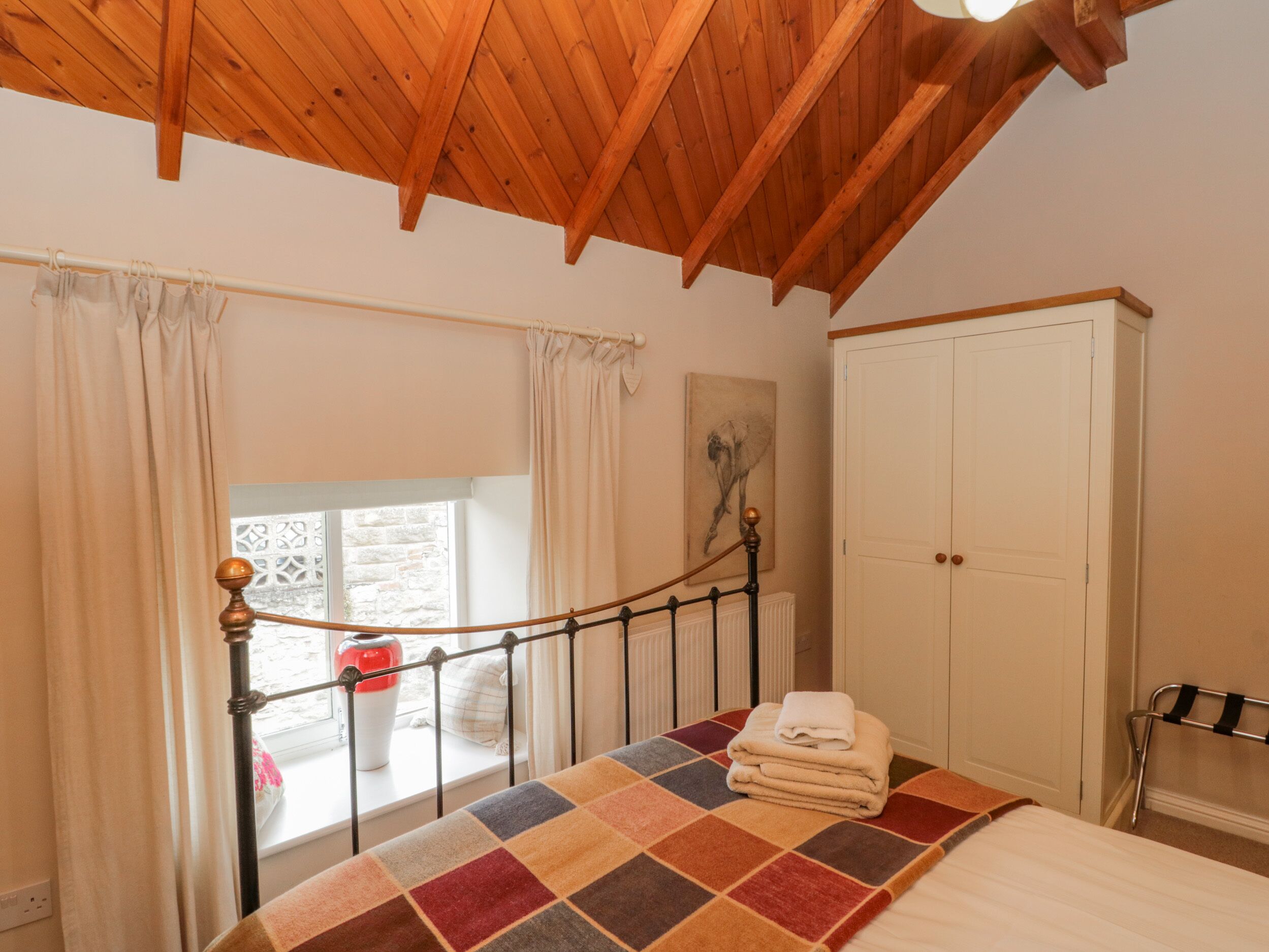 A bedroom with a wooden ceiling, a bed with a colorful quilt, a window with curtains, and a white wardrobe.