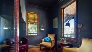 Una habitación con paredes oscuras, suelo de madera, un sillón de cuero rojo, un sillón beige con un cojín azul, una mesa de madera con una planta en maceta y una ventana con contraventanas de madera.