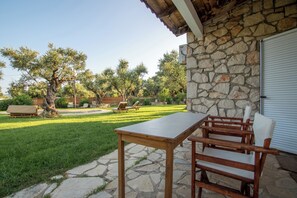 A patio with a stone table and wooden chairs, a stone wall, and a view of a grassy yard with trees and a building in the background. - Il Greco   (Zakynthos)