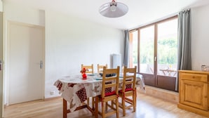 A dining area with a table set for four, wooden chairs, a ceiling light, and a view through a large window. - L'Ourson des Myrtilles (La Bresse)