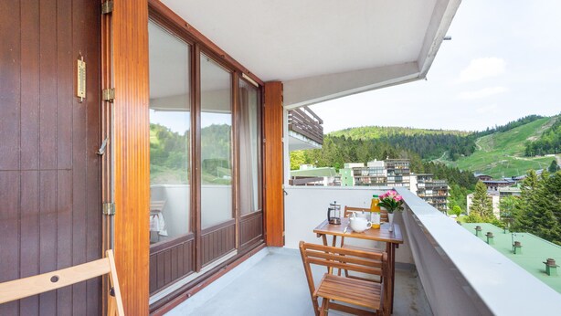 A balcony with a wooden table and chairs, a view of a green valley, and a building in the background. - L'Ourson des Myrtilles (La Bresse)