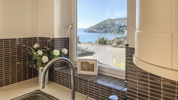 A bathroom with a sink, a window with a view of a coastal town, and a vase with white flowers.
