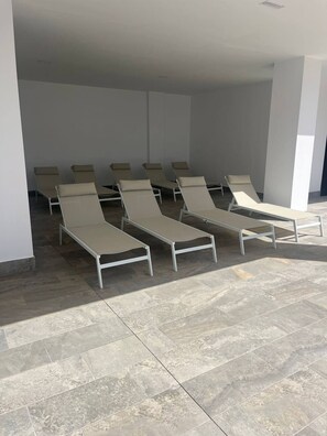 A row of sun loungers with metal frames and beige cushions, arranged in an indoor setting with tiled flooring and white walls.