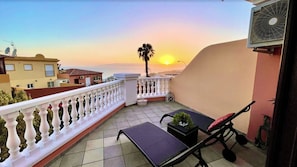 Ein Balkon mit Blick auf den Sonnenuntergang, eine Palme und ein Sessel mit Kissen.