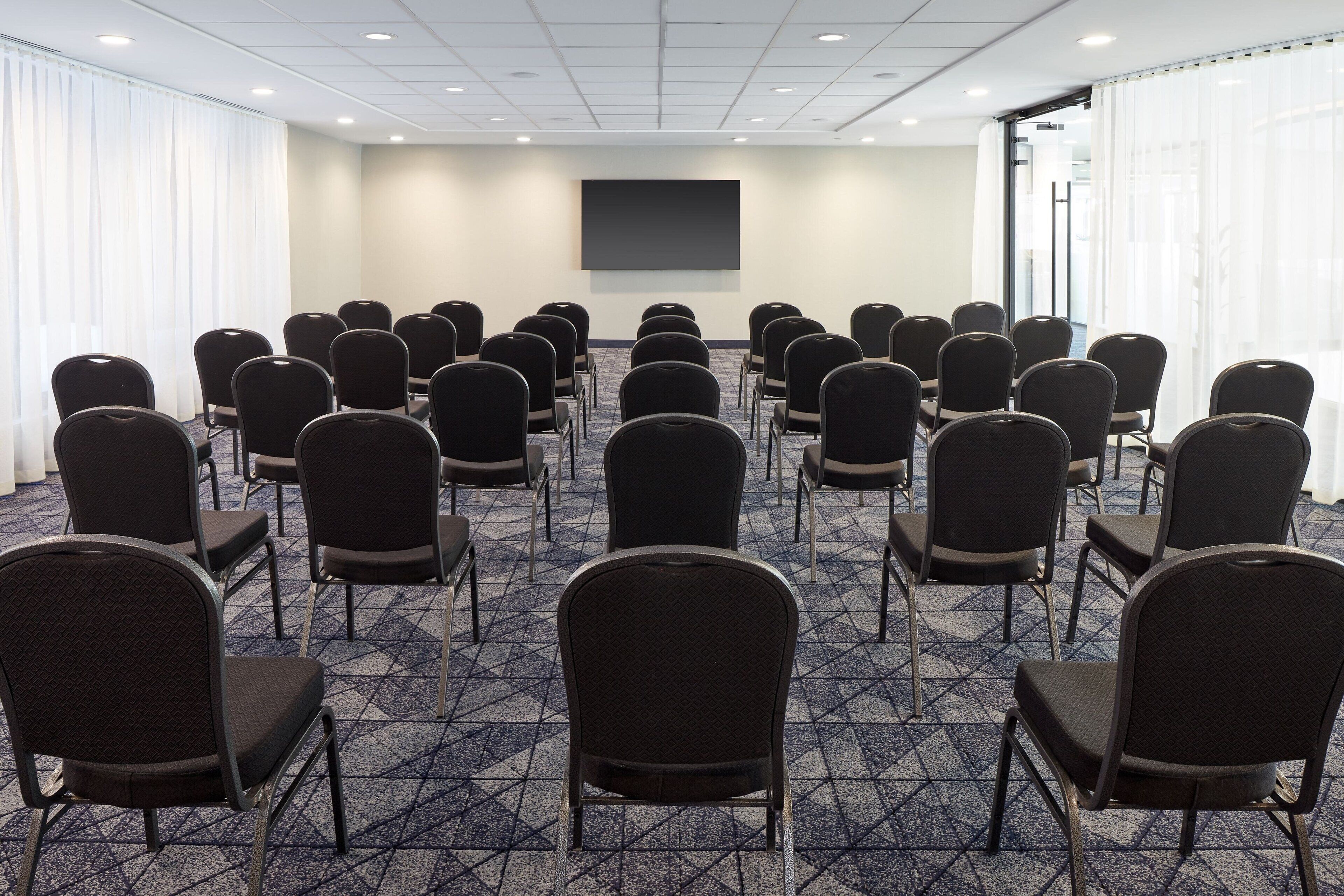 A conference room with rows of chairs facing a large screen, carpeted floor, and windows with curtains.