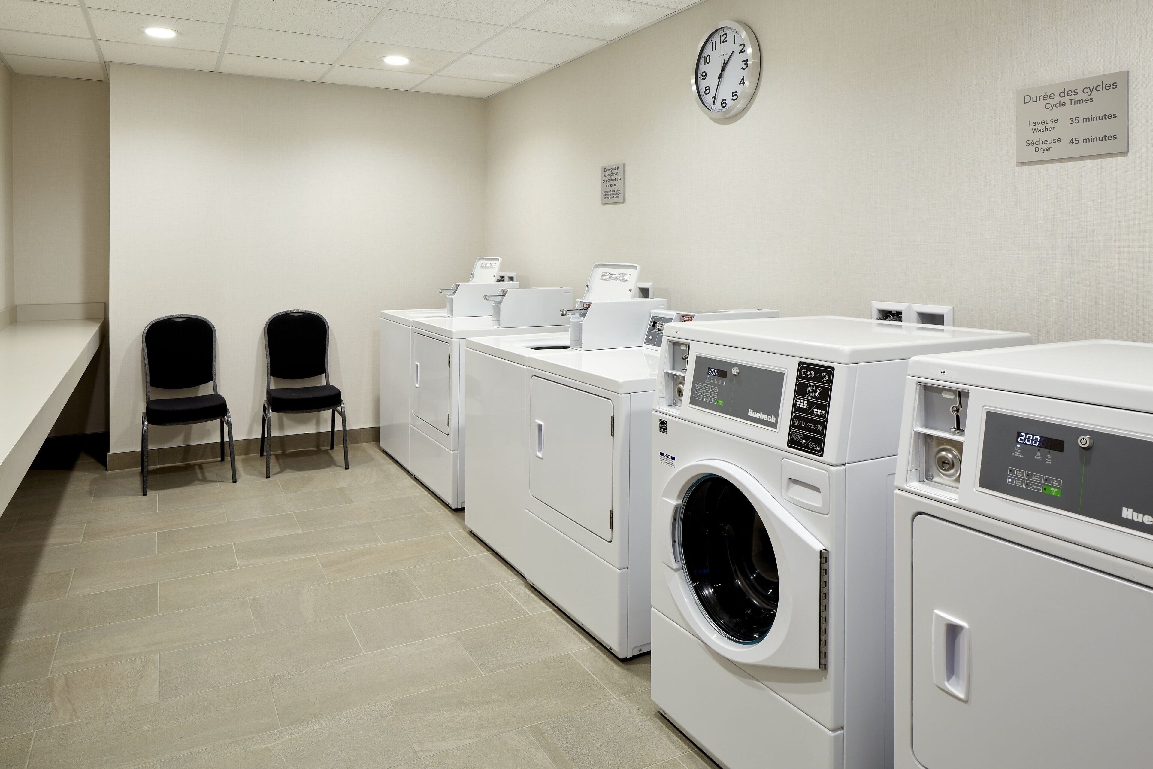 A laundry room with four washing machines, two black chairs, a clock, and a sign indicating cycle times.