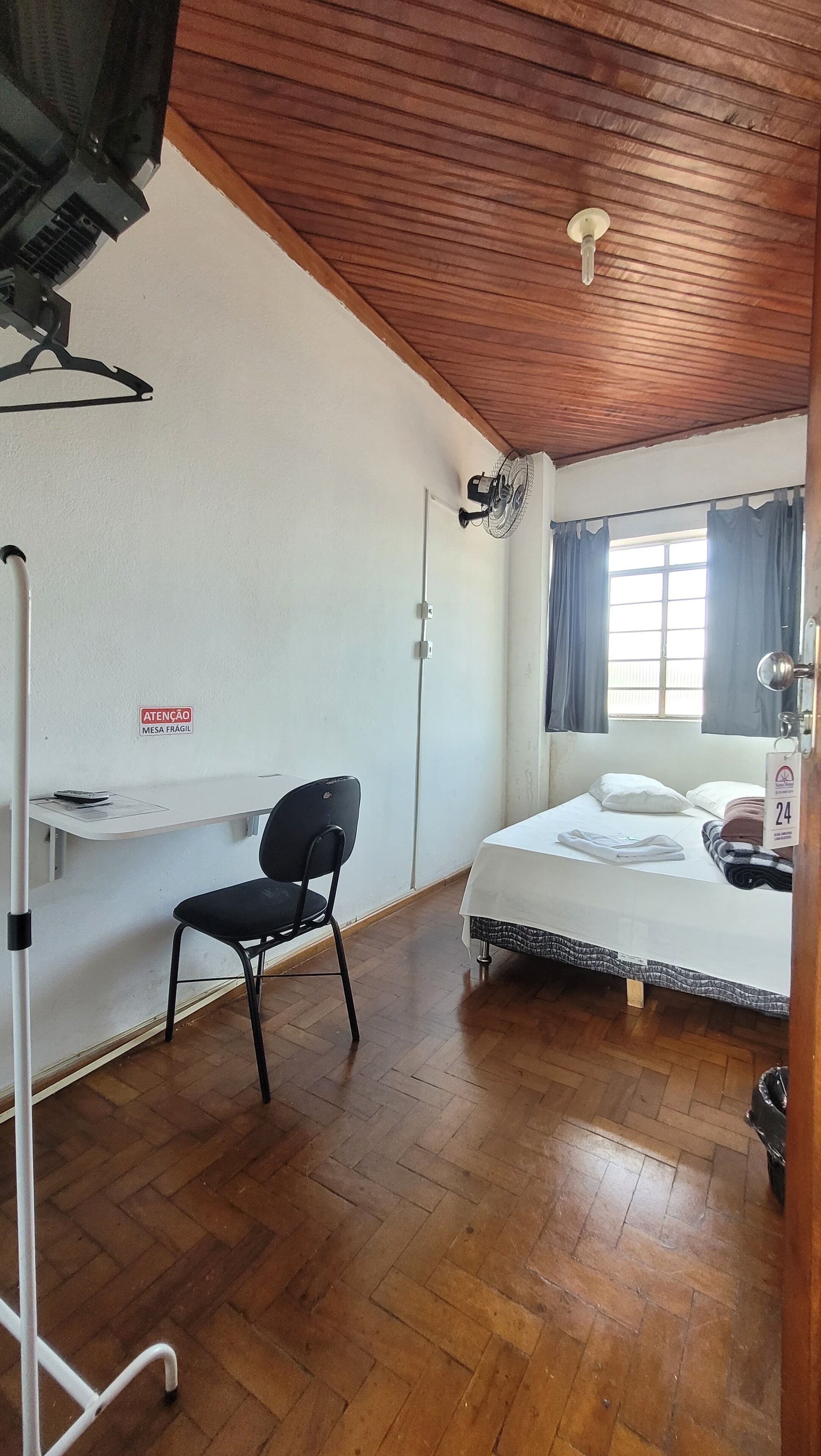 Une chambre d’hôtel équipée d’un lit, d’un bureau, d’une chaise, d’un ventilateur de plafond et d’une fenêtre avec des rideaux.