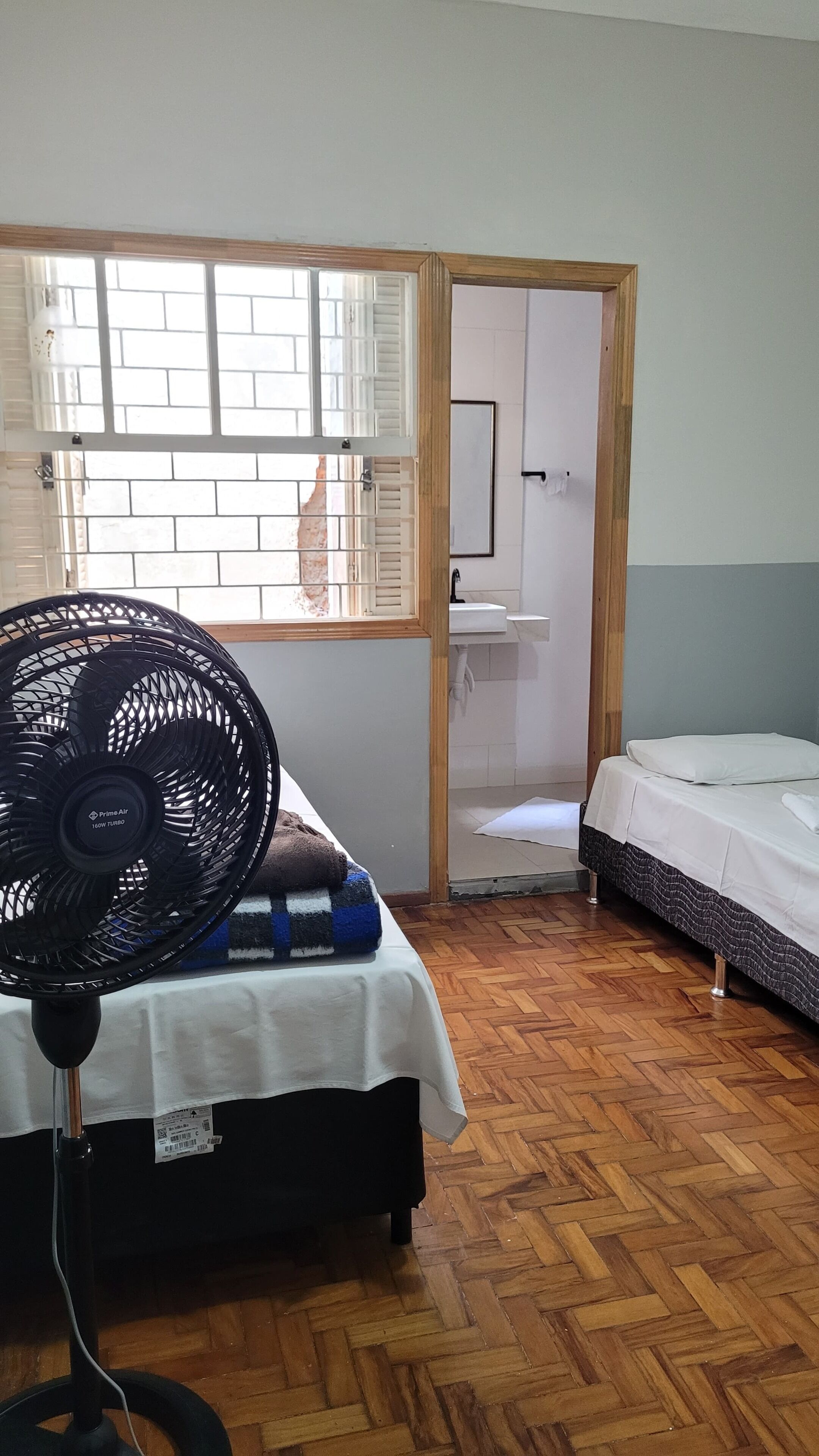 Una habitación con cama, ventilador, ventana con contraventanas y puerta que da a un baño.