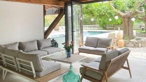 Une pièce spacieuse avec un plafond en bois et de grandes fenêtres, un ensemble de canapés gris, une table basse, et une vue sur une piscine et la verdure environnante.