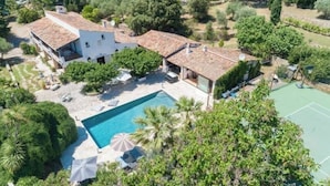 Une maison à deux étages avec une piscine, entourée d’une végétation luxuriante et dotée d’un court de tennis.