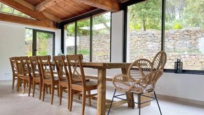 Un espace repas avec une table et des chaises en bois, de grandes fenêtres donnant sur un mur en pierre et un plafond en bois.