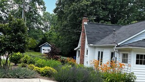 Une maison blanche avec une cheminée en briques, entourée d’un jardin bien entretenu où poussent différentes plantes et fleurs.