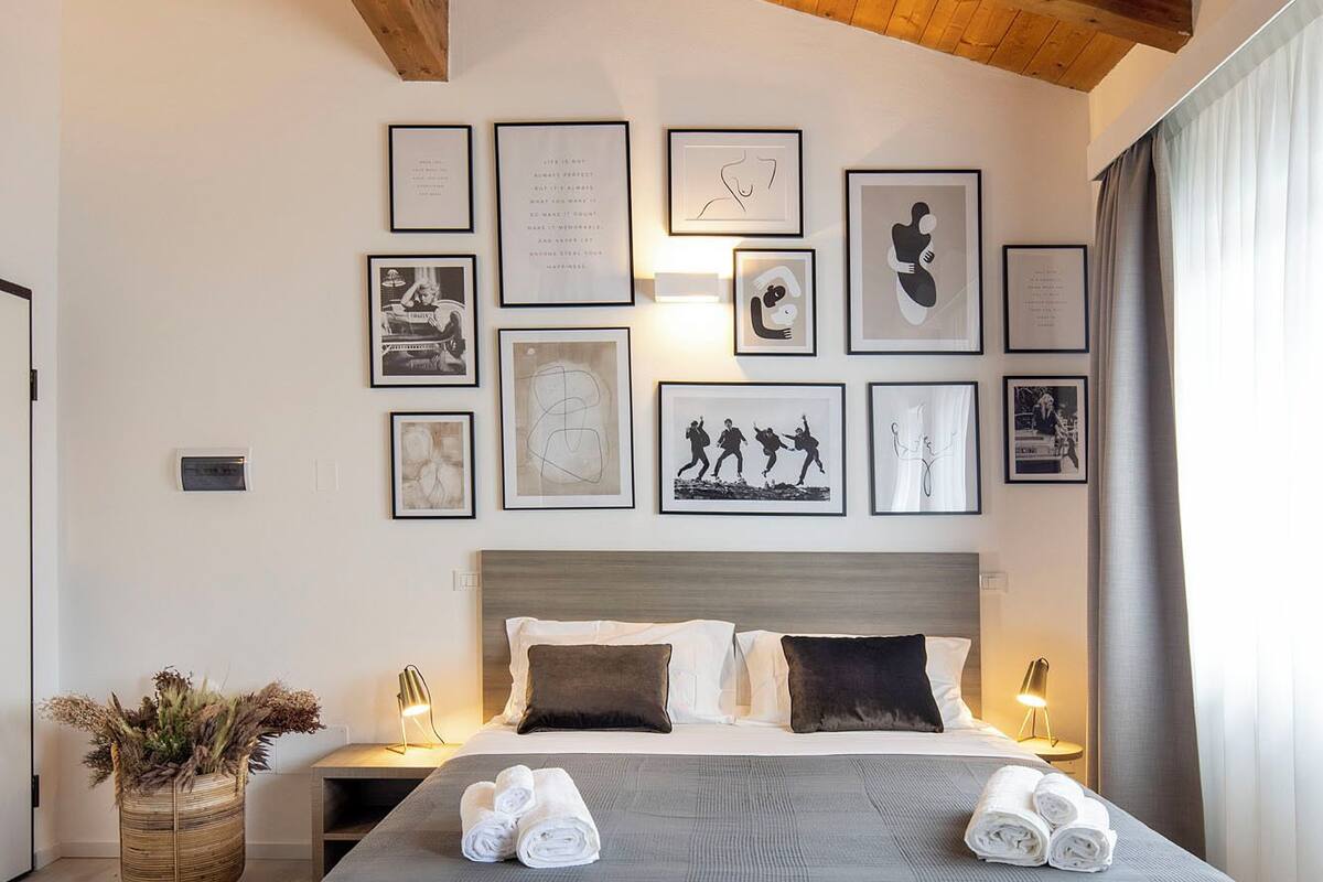 A modern bedroom with a wooden ceiling, a large bed with grey bedding, and multiple framed artworks on the wall.