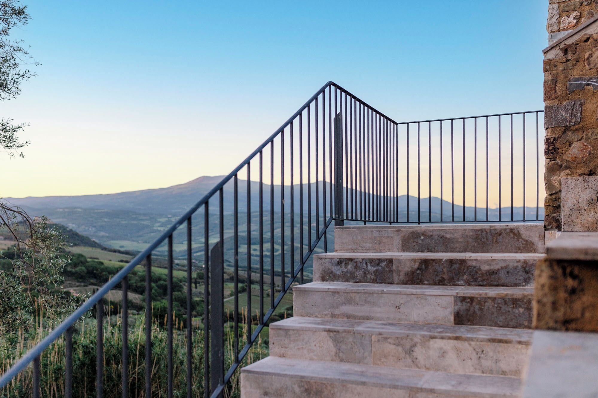 A staircase with a metal railing leading to a stone building, overlooking a mountainous landscape.