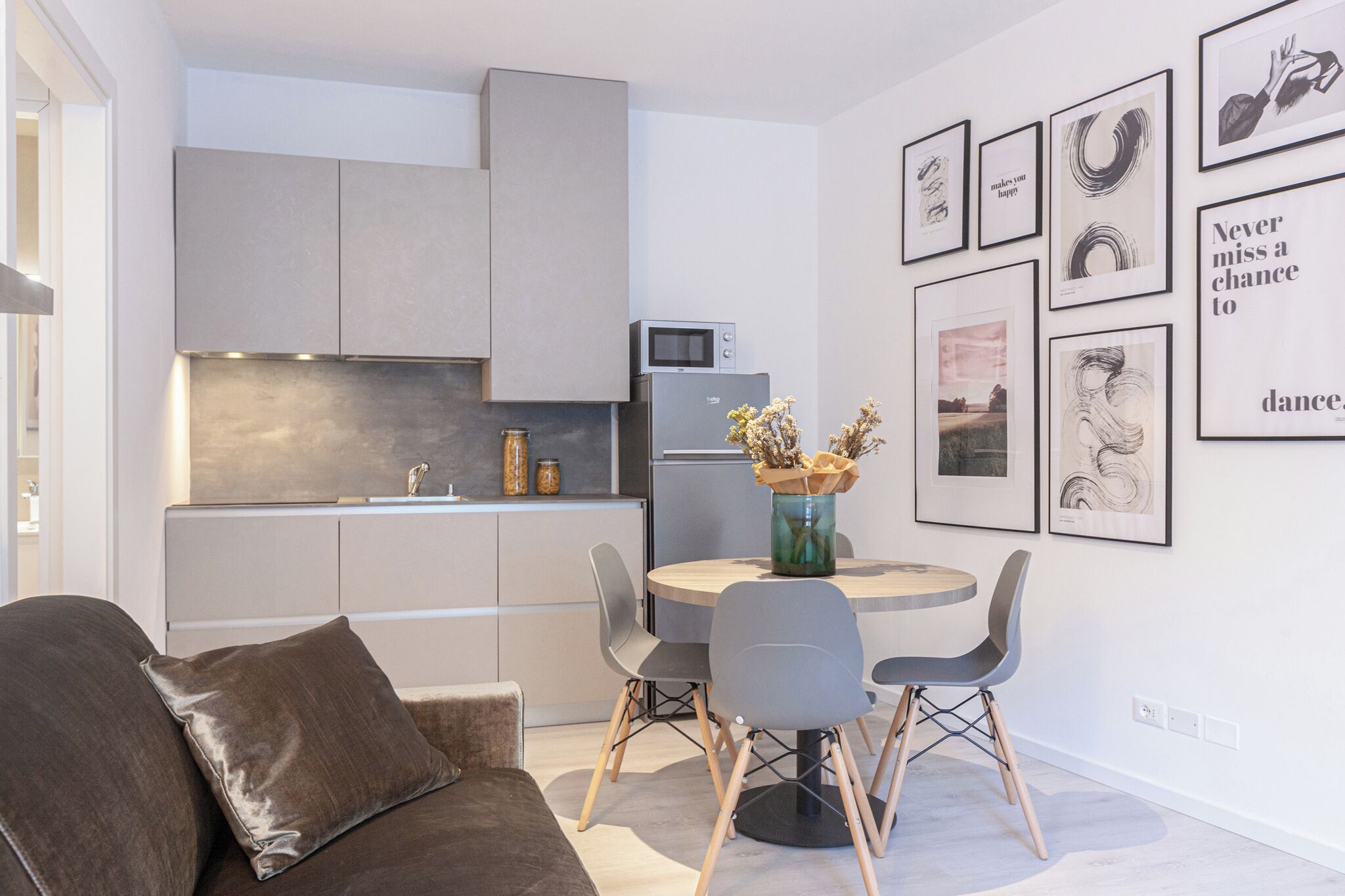 A modern kitchen with grey cabinets, a built-in microwave, and a refrigerator. There is a dining table with four chairs and a vase with flowers. The walls are adorned with framed artwork.