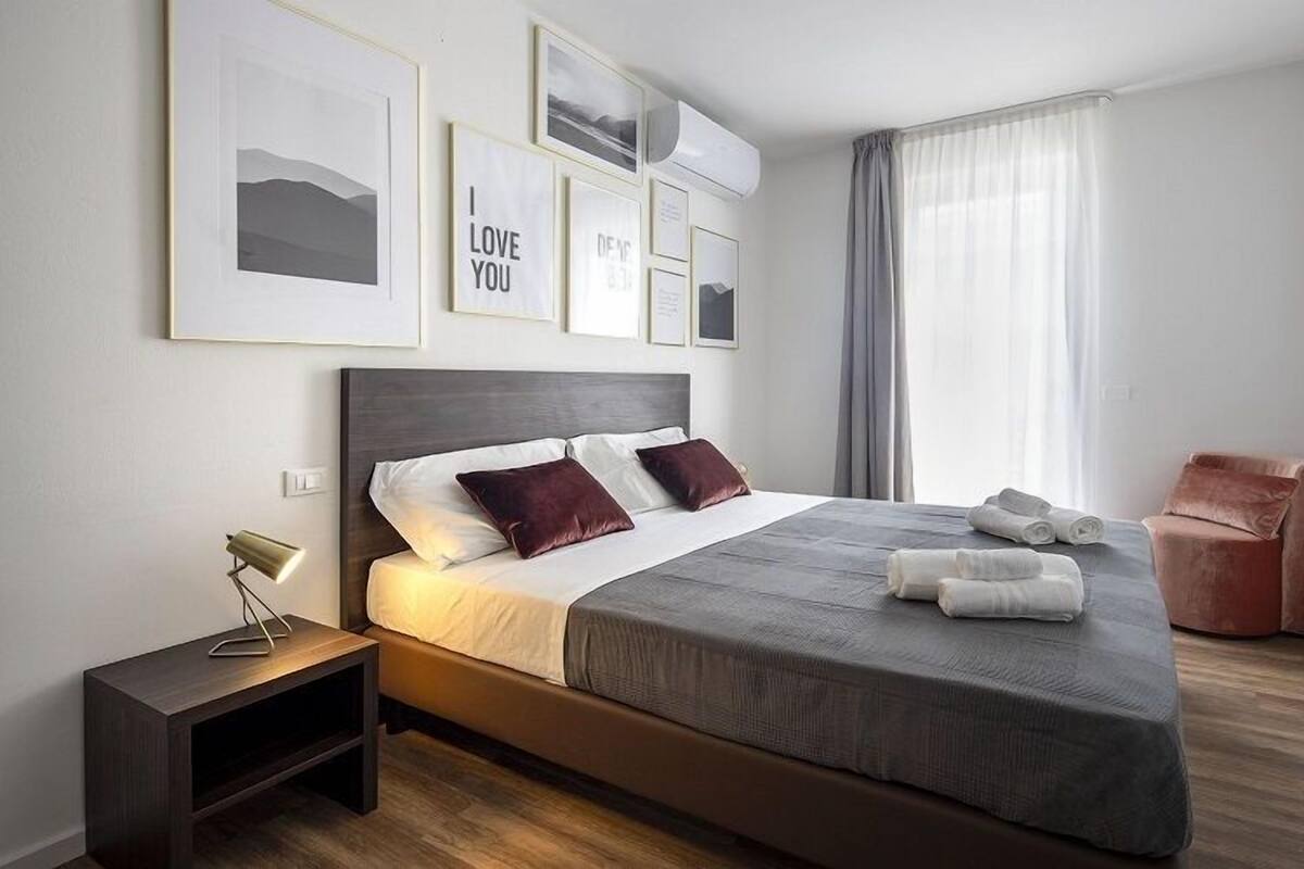 A modern hotel room with a large bed, a bedside table, a chair, and a window with curtains.