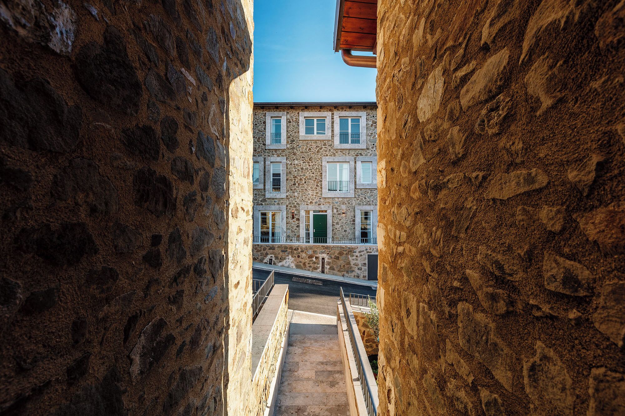 Un camí de pedra estret que mena a un edifici amb diverses finestres i una porta verda.
