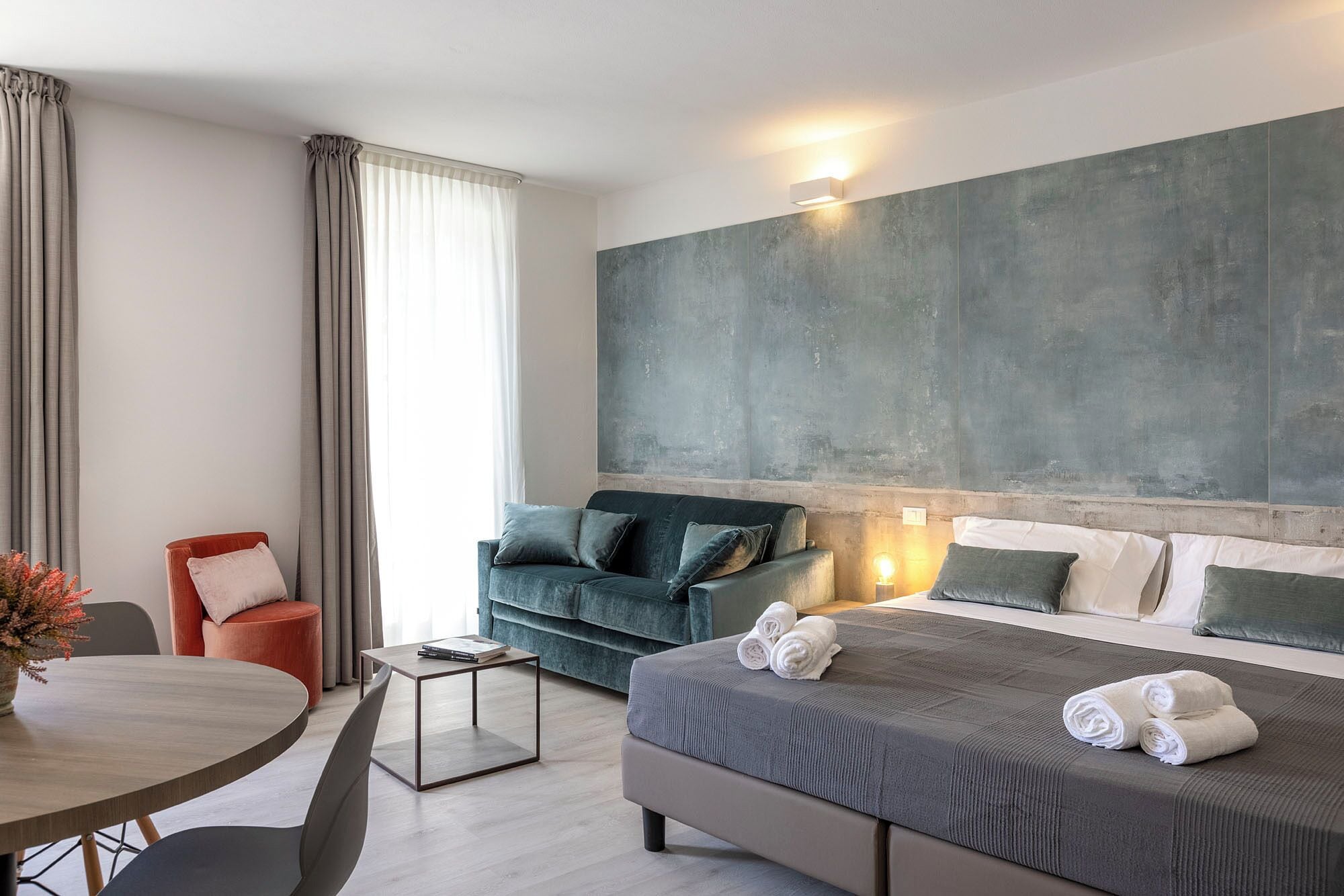 A modern hotel room with a large bed, a sofa, a dining table, and a large abstract painting on the wall.