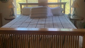 A bed with a striped bedspread, two white pillows, and a wooden headboard. There are two bedside tables with lamps, a hanging plant, and a window with a view of trees. - Ranch in equestrian community (Murrieta)