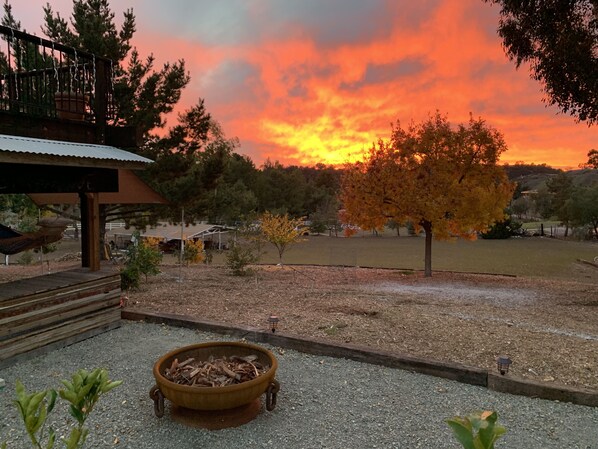 A patio area with a fire pit, a covered seating area, and a view of a sunset over a grassy field.