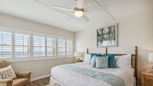 Une chambre dotée d’un grand lit, d’un ventilateur de plafond, d’une fenêtre avec des stores, d’une table de chevet avec une lampe et d’une œuvre d’art accrochée au mur.