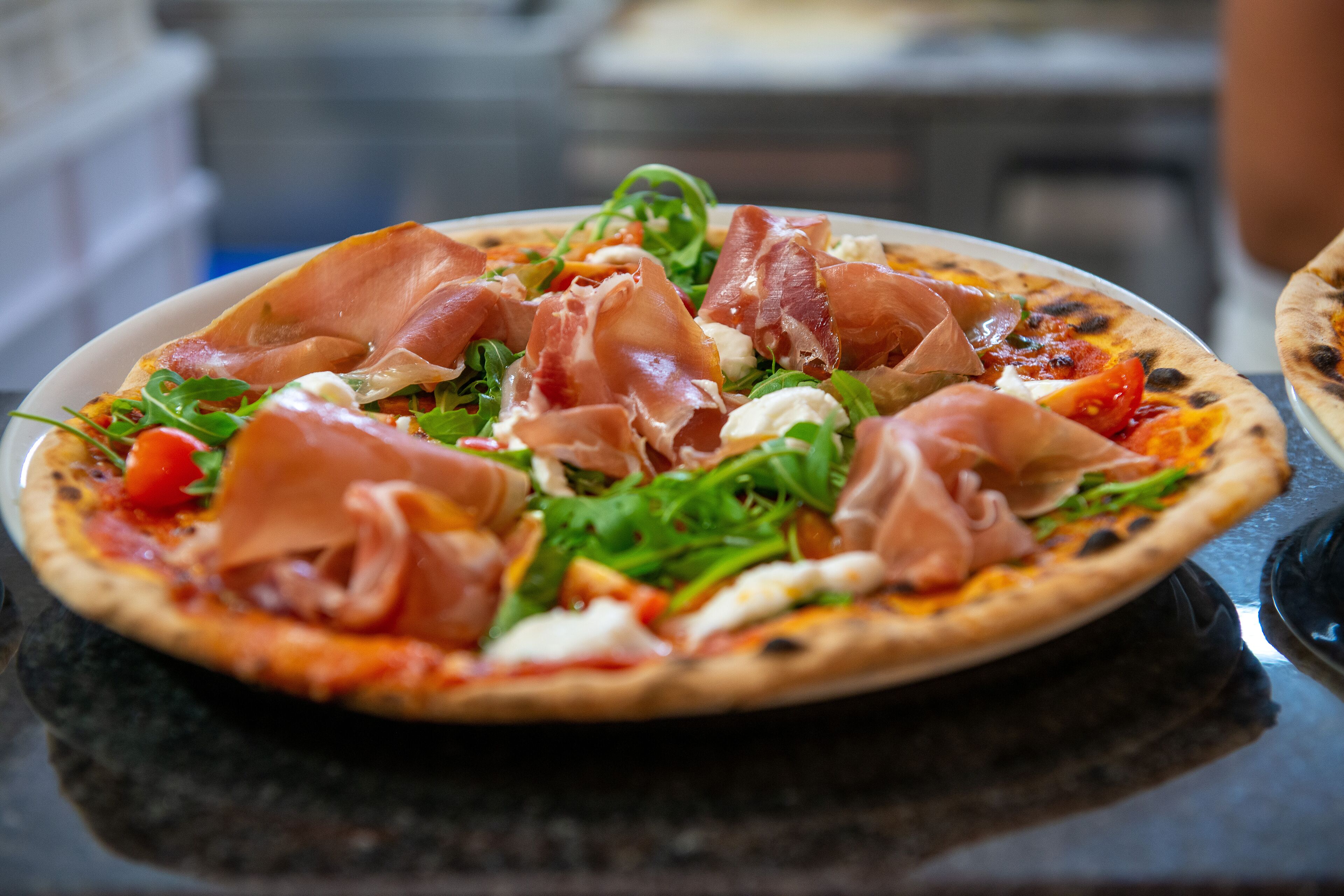 Pizza mit Schinken und Grünzeug auf einem schwarzen Tisch.