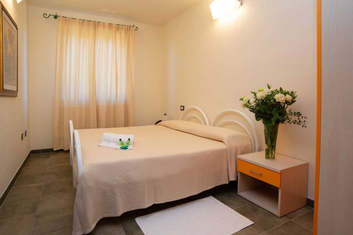 Una habitación de hotel con cama, mesita de noche, un jarrón con flores y ventana con cortinas.