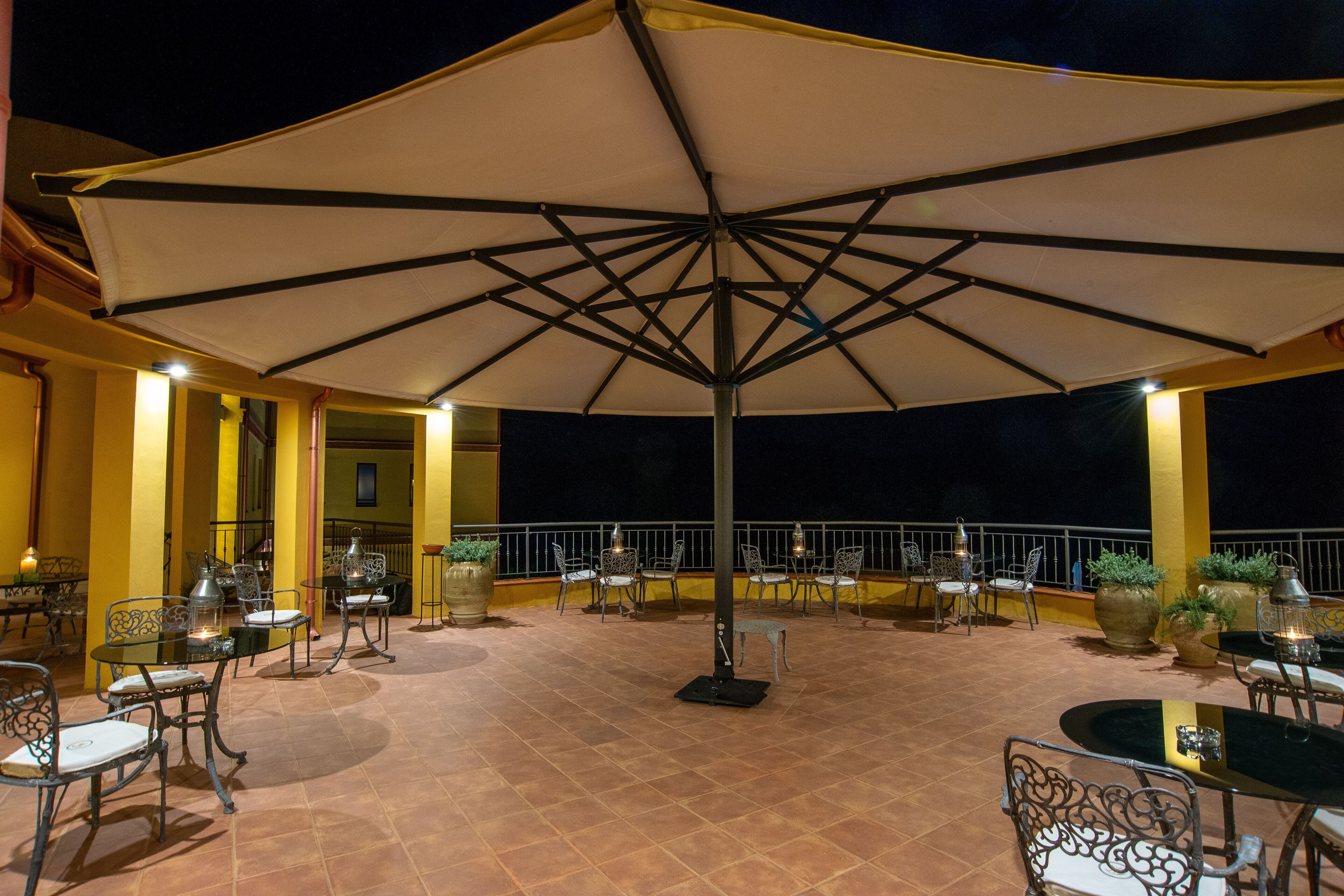 Una terrazza esterna con tavoli e sedie sotto un ombrellone grande, circondata da pareti gialle e piante in vaso.