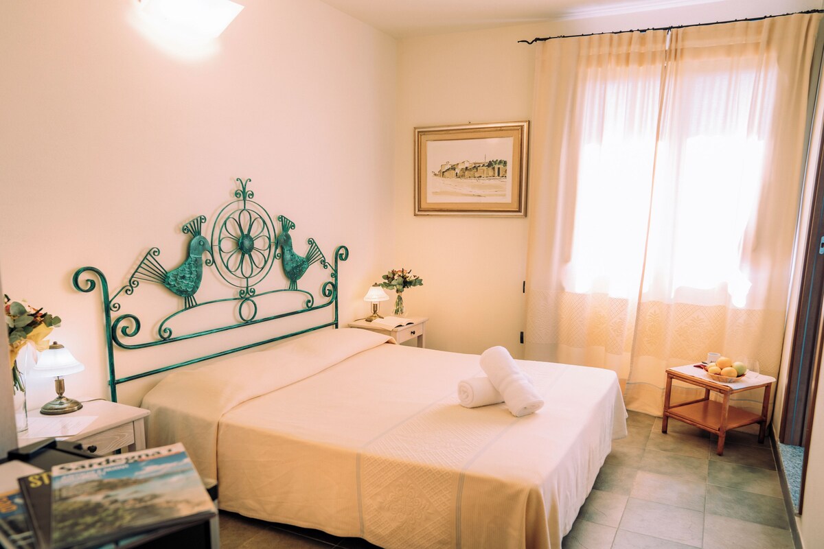 Habitación de hotel impecable con una cama, una mesita de noche con lámpara, una pequeña mesa con frutas y un cuadro en la pared.
