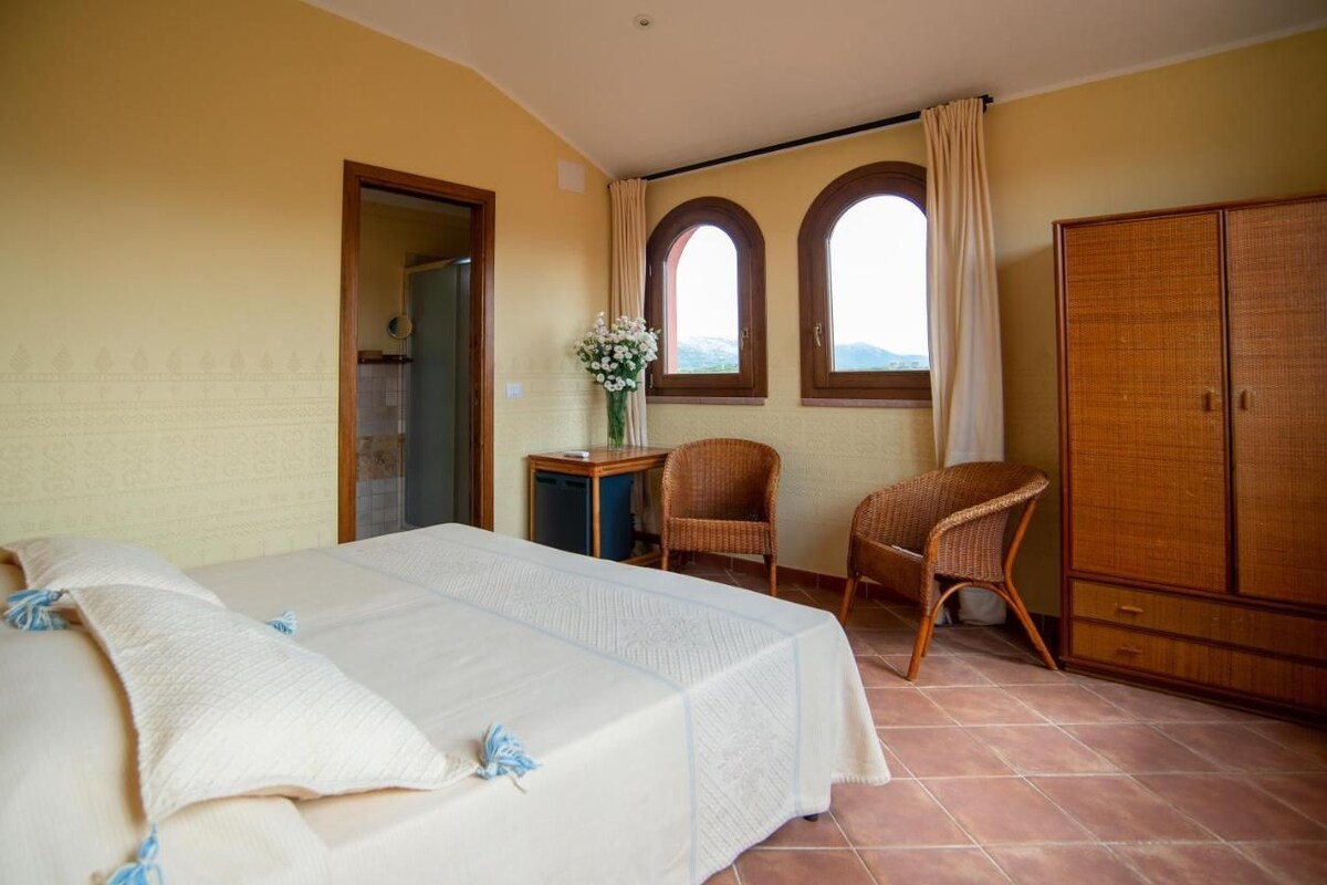 Un dormitorio con una cama, dos sillones de mimbre, una mesita con un jarrón y un armario.